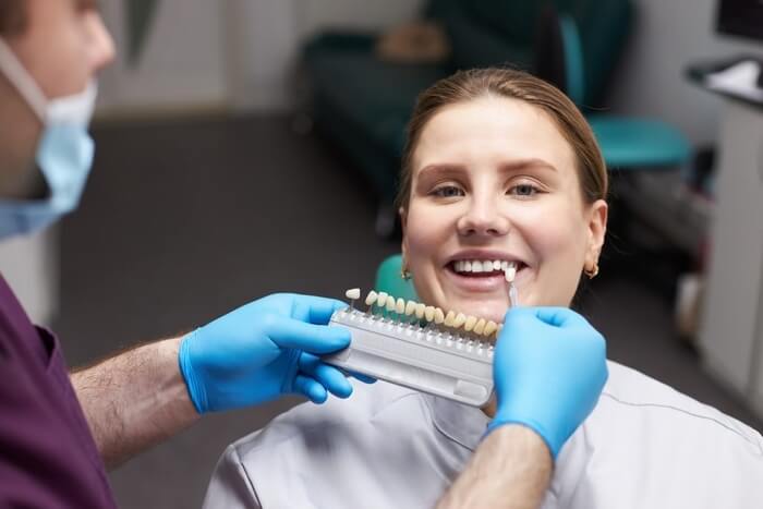 Close-up smiling pretty woman patient in dentist's seat, looking at camera while doctor orthodontist, choosing the shade of dental veneer according to color chart. Aesthetic dentistry. Dental practice